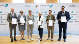 Vier Frauen und zwei Männer stehen mit Dokumenten in ihren Händen vor einer Stellwand mit der Aufschrift "Bildungsland NRW".