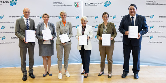 Four women and two men stand with documents in their hands in front of a display wall with the inscription "Bildungsland NRW".