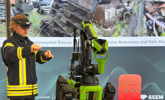 Foto: Eine Person in Feuerwehrkleidung trägt eine VR-Brille und steuert einen grünen Roboter mit Greifarm in einer Innenumgebung.