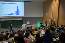 Ein voll besetzter Hörsaal von der oberen Reihe aus. Vorne steht Andreas von Bechtholsheim, ein Mann mit grauen Haaren und einem hellblauen Hemd, mit Mikrofon und spricht. Links von ihm steht Rektor Manfred Bayer und hört zu.