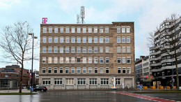 Frontansicht von einem Bürogebäude mit einem Telekom-Logo auf dem Dach. Drumherum sieht man die Straßen und umliegende Gebäude.