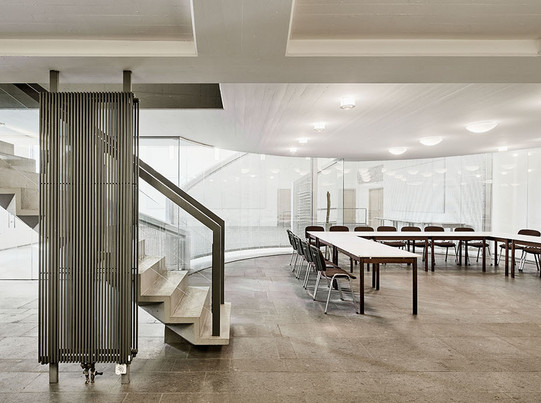 Auf dem Foto sieht man einen großen, hellen Raum mit einer Treppe, Tischen und Stühlen.