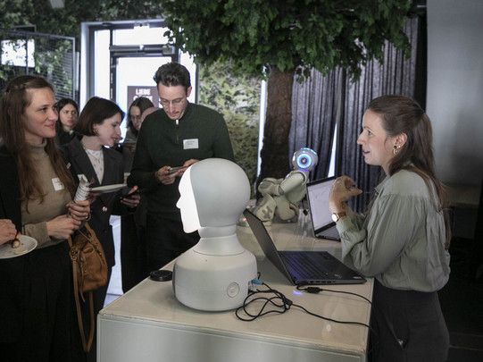 Mehrere Personen stehen an einem Messetresen und betrachten einen humanoiden Roboterkopf, daneben ein kleiner Roboter und zwei geöffnete Laptops.