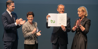 A smiling Donald Tusk holding his Honorary Doctorate certificate into the camera. Professor Schuck, Rita Süssmuth and Rector Ursula Gather stand next to him and applaud