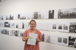 Eine Frau steht vor einer weißen Wand an der mehrere Bilder einer Kunstausstellung hängen und hält eine Urkunde hoch.