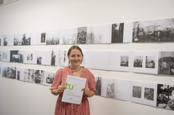 Eine Frau steht vor einer weißen Wand an der mehrere Bilder einer Kunstausstellung hängen und hält eine Urkunde hoch.