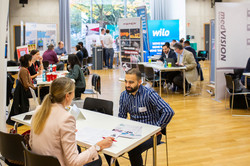 Gespräche auf der Messe In einem großen Raum stehen viele Tische, an denen Menschen Gespräche führen