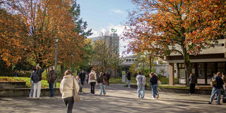 Foto: Ein Foto vom Campus der TU Dortmund im Herbst. Auf dem Foto sind Studierende, Food Fakultät und der Mathetower mit dem TU Logo zu sehen.