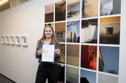 Eine Studentin mit Urkunde steht vor 20 Einzelfotografien an der Wand.