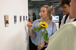 Eine Person, die auf Polaroid-Fotos zeigt und etwas erklärt. Sie hält eine Sonnenblume in der Hand.