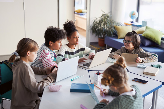 Mehrere Kinder sitzen im Kreis um einen Tisch. Zwei Kinder haben einen Laptop, ein weiteres Kind ein Tablet vor sich aufgestellt. Eines der Kinder schreibt mit einem Stift in ein Heft.