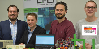 Haptical Gruppenfoto von vier Männern, die vor einem Tisch stehen auf dem ein Laptop und Technik-Bauteile liegen.