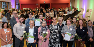 A group photo of various people with Prof. Tessa Flatten in front of a packed hall. Three people hold certificates in their hands. A man with gray hair and glasses holds a white rose next to the certificate. A woman in a gray-black hijab with a geometric pattern and a woman with glasses and short brown hair each hold a white rose.