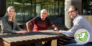 Zwei Männer und eine Frau sitzen draußen an einem Tisch auf dem Campus der TU Dortmund.