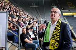 Ein Mann mit Anzug und Fanschal steht vor einer gefüllten Tribüne im Stadion.