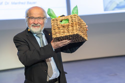 Vortragsreihe „Initialzündung“ Ein Mann im Anzug hält einen Präsentkorb in die Luft.