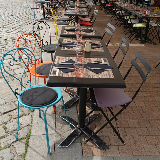 Café Auf einem Bürgersteig stehen drei kleine Tische mit bunten Stühlen drum herum.