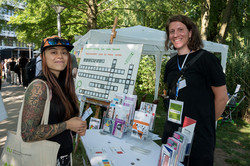 Eine junge Frau und eine Mitarbeiterin stehen an einem Informationsstand zur Antidiskriminierungsarbeit und lächeln in die Kamera.