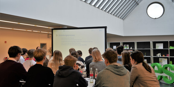 Junior-Innovation-Day Schüler*innen sitzen an Tischen in einem Raum und schauen auf einen großen Bildschirm.