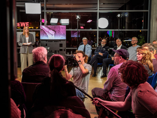 Foto: Eine Person spricht vor einem sitzenden Publikum in einem beleuchteten Studio.