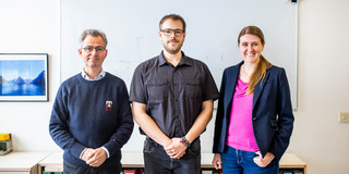 A portrait of two men and a woman in front of a whiteboard with formulas