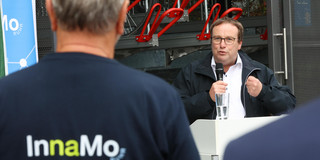 NRW-Verkehrsminister spricht in ein Mikrophon. Im Vordergrund ist der Rücken eines Mannes zu sehen, der ein T-Shirt mit der Aufschrift "InnaMo Fahrradhub" trägt.