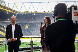 Ein Kameramann filmt Christoph Edeler (links) und eine Frau mit Mikrofon in der Hand (rechts). Hinter ihnen sieht man das Innere des Stadions.