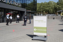 A signpost to the exams stands in front of a large building with a glass front. In Front of the entrance five people are standing in a circle.