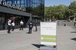 Klausur in den Westfalenhallen Ein Wegweiser-Schild zu den Klausuren steht vor einem großen Gebäude mit Glasfront, vor dessen Eingangstür stehen fünf Personen im Kreis.
