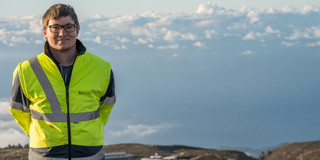 A young man in a yellow TU-Dortmund high-visibility vest stands elevated on a mountain, behind him a hilly panorama and cloudy sky.