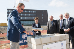 Several people are standing around a brick cube and want to sink a time capsule. A man is filling the cube with a putty knife and putty.