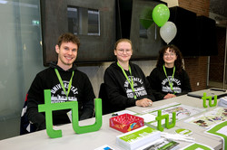 The picture shows a row of white tables in the foreground with lots of information material about TU Dortmund University. Behind the row of tables sit three people in their black university hoodies.