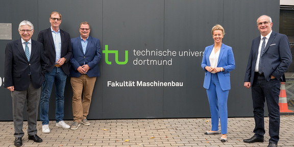 Ina Brandes, Prof. Manfred Bayer, Prof. Gerhard Schembecker, Albrecht Ehlers und Prof. A. Erman Tekkaya stehen vor einer Mauer, auf der das Logo der TU Dortmund und der Schriftzug "Fakultät Maschinenbau" zu sehen ist.
