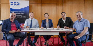 Fünf Personen sitzen hinter einem Tisch. Vor ihnen liegen Dokumente, die vier der fünf Personen unterschreiben.