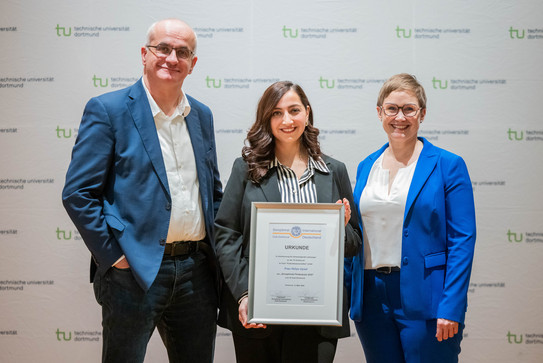 Von links nach rechts: Rektor Prof. Manfred Bayer, Lehramtsstudentin Hülya Uysal und Daniela Fischer vom Soroptimist International Club Dortmund vor einem weißen Backdrop, auf dem mehrfach das Logo der TU Dortmund steht. Die Studentin hält eine Urkunde in den Händen.