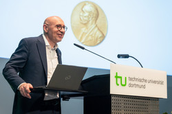 Foto: Eine Person steht am Pult der Technischen Universität Dortmund mit einem Laptop, während im Hintergrund eine Medaille auf der Leinwand zu sehen ist.