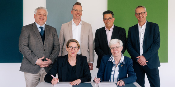Auf dem Foto sieht man insgesamt sechs Personen in die Kamera lächeln. Zwei von den sechs Personen sitzen an einem Tisch und unterzeichnen ein Dokument.