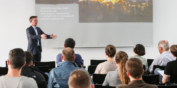 Ein Mann steht vor einer Wand, auf die eine Präsentation projiziert wird. Vor ihm sitzen mehrere Personen auf Stühlen.