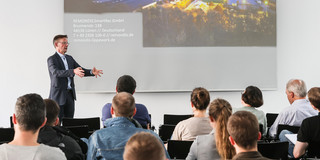 Ein Mann steht vor einer Wand, auf die eine Präsentation projiziert wird. Vor ihm sitzen mehrere Personen auf Stühlen.