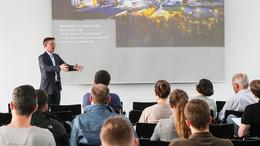 Ein Mann steht vor einer Wand, auf die eine Präsentation projiziert wird. Vor ihm sitzen mehrere Personen auf Stühlen.