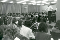 A conductor stands on a pedestal, people sit in front of him and sing.