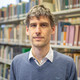 Portrait von Giulio Mattioli. Im Hintergrund ist ein großes Bücherregal der Bibliothek zu sehen.