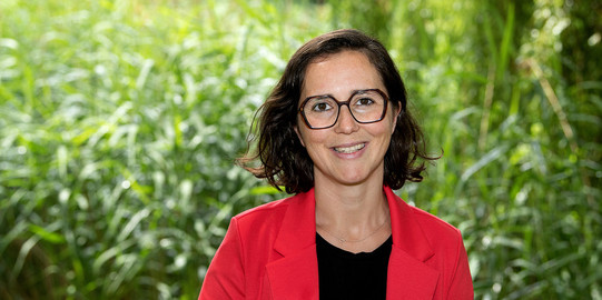 Porträtfoto von Prof. Liane Bächler: Prof. Bächler trägt ein schwarzes Oberteil, einen roten Blazer und eine dunkle Brille. Sie steht vor Pflanzen auf dem Campus der TU Dortmund.