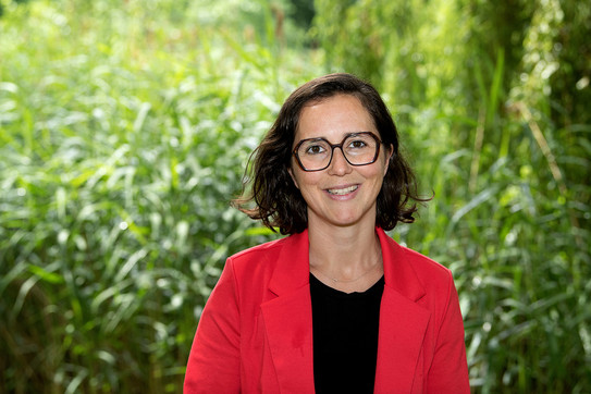 Porträtfoto von Prof. Liane Bächler: Prof. Bächler trägt ein schwarzes Oberteil, einen roten Blazer und eine dunkle Brille. Sie steht vor Pflanzen auf dem Campus der TU Dortmund.
