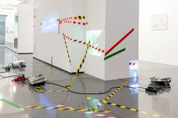 Ausstellung „Laufende Beobachtungen“ In einer Ausstellung kleben verschiedene bunte Klebestreifen an einer weißen Wand. Davor steht ein Beamer und es liegen Kabel auf dem Boden.