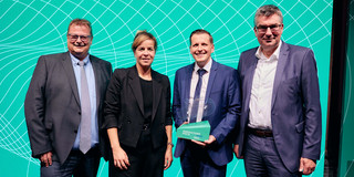 Drei Männer und eine Frau posieren vor einer türkisfarbenen Wand. Sie tragen Anzüge und der dritte Mann von links hält eine Auszeichnung mit der Inschrift "Innovationspreis NRW 2023" in den Händen.