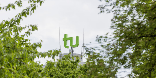 Das grüne TU-Logo umrahmt von Bäumen.