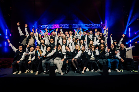 Gruppenfoto: Jubelnde Gruppe von Menschen in Teamjacken vor bunter Bühne der BRYCK Startup Alliance.
