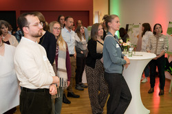 In the photo, you can see several people standing at high tables, watching and listening to presentations.