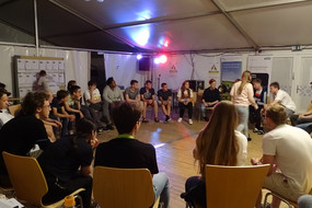 Do-camp-ing Teilnehmerinnen und Teilnehmer sitzen gemeinsam in großer Runde im Zelt zusammen.